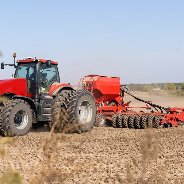 Un grand tracteur rouge avec des roues arrière jumelées tirant un semoir dans un champ labouré sous un ciel bleu clair.