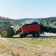A green tractor towing a large red hay baler across a field, processing straw under a bright sky.