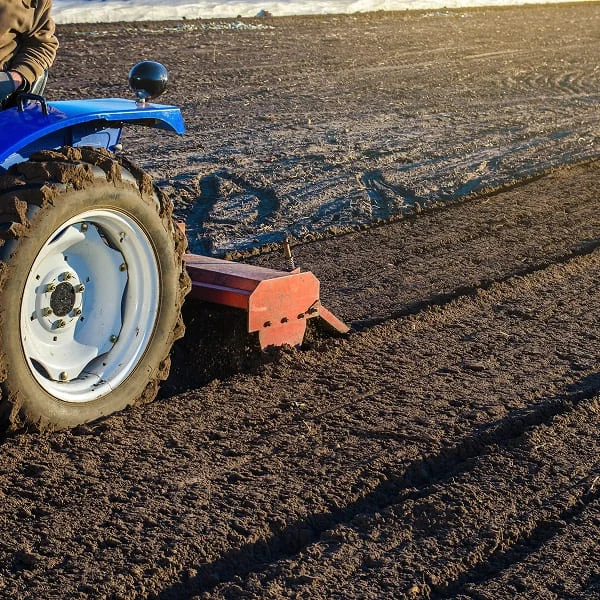 A man operates a tractor, plowing a field under clear blue skies, preparing the soil for planting.